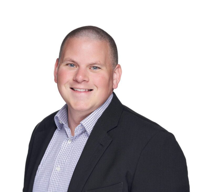 A man with short hair is smiling, wearing a black blazer over a checkered button-up shirt, posed against a plain white background.