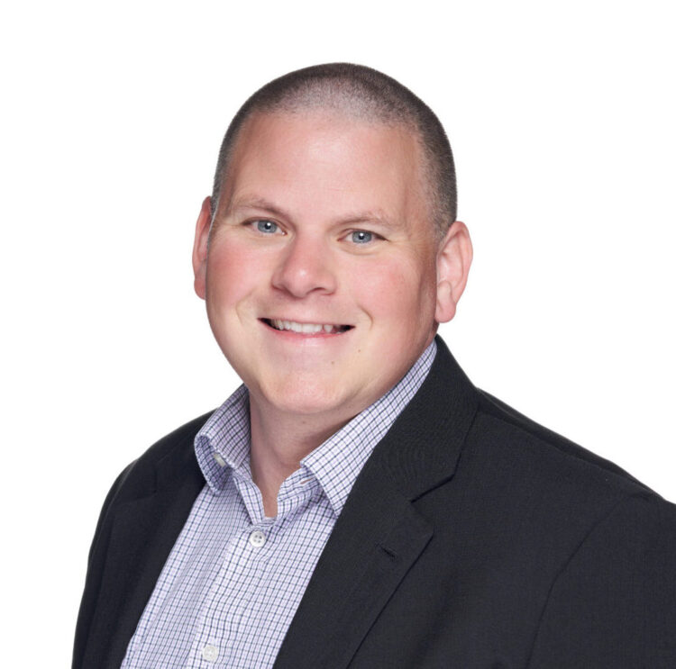 A man with short hair is smiling, wearing a black blazer over a checkered button-up shirt, posed against a plain white background.