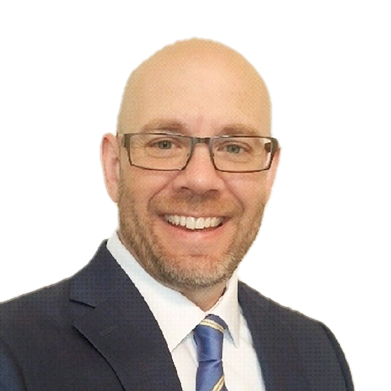 A man with glasses, a beard, and a bald head is smiling. He is wearing a dark suit, white shirt, and striped tie, posed against a plain white background.