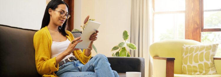 A woman sits on a couch using her tablet to explore content portal benefits, wearing glasses and a yellow cardigan. A coffee cup rests on the table as sunlight streams through the window.