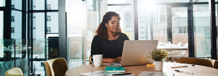 A woman sits at a round table in a modern office, working on her laptop with a coffee mug, notebook, and small plant nearby as she researches how to choose a cloud provider.