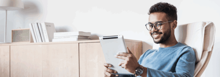 A man with glasses sits in a chair, smiling while using a tablet to explore an access portal. A wooden cabinet with books and a speaker is in the background.