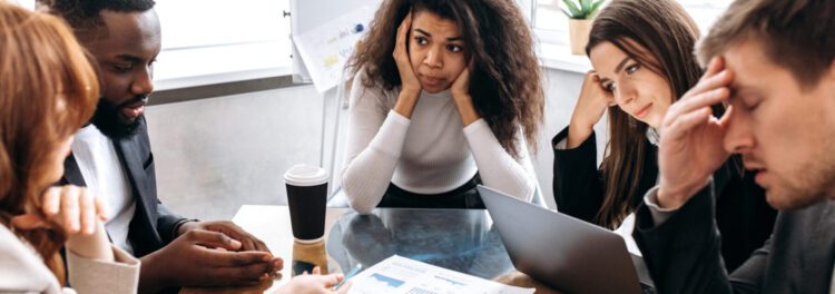 Five people sit around a table with papers and a laptop, appearing stressed or frustrated during a meeting—perhaps discussing why digital transformations fail.