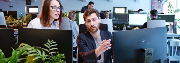 Two colleagues work together at a computer in a modern office with other employees at desks in the background, demonstrating why managed services are essential for seamless collaboration and productivity.