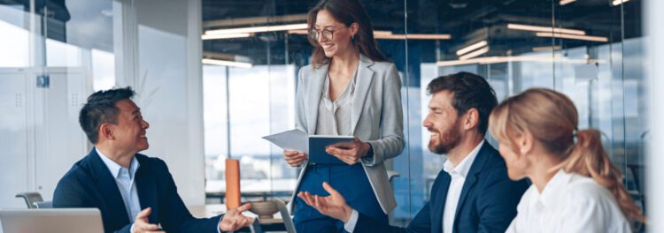 Four business professionals in formal attire sit and stand around a conference table, engaged in discussion about finding the right digital transformation partner in a modern office setting with large windows.