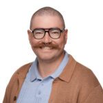 A person with short hair, glasses, a mustache, and a septum piercing, wearing a light blue collared shirt and a brown jacket, smiles against a white background.