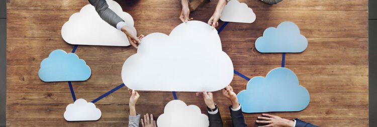 Several people’s hands holding cutout cloud shapes over a wooden table, with lines connecting the clouds—symbolizing a Cloud-First Strategy and network connection.