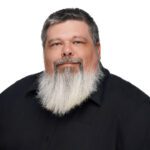 A man with short dark hair, a long gray and white beard, and a black collared shirt poses against a plain white background.