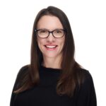 Smiling woman with long brown hair, wearing glasses and a black top, posed against a plain white background.