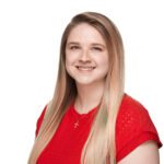 Young woman with long blonde hair wearing a red top and a cross necklace, smiling against a plain white background.