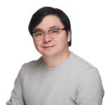A person with short dark hair and glasses wearing a light gray long-sleeve shirt poses against a plain white background.