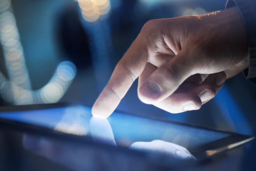 A close-up of a hand touching the screen of a tablet device, with blue and white lights blurred in the background.