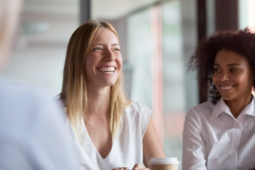 Two women in white shirts sit at a table, smiling and talking, with a coffee cup in the foreground and large windows in the background.