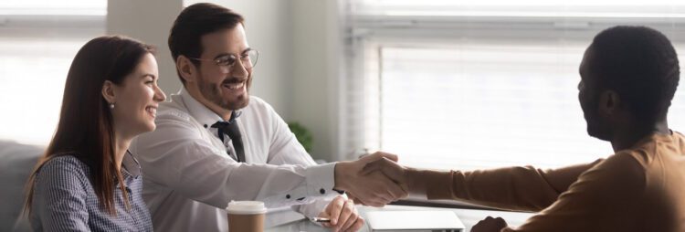 A man and woman sit at a table smiling while the man shakes hands with another man across from them in an office setting, discussing RPA Insurance Use Cases.