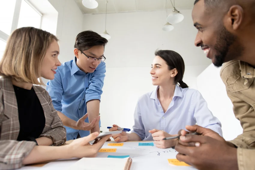 Four people sit and stand around a desk, engaged in discussion with notebooks, pens, and smartphones in a well-lit office setting.