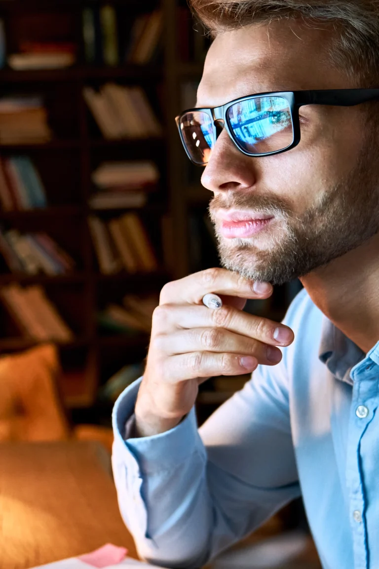 Man in glasses and blue shirt sits indoors, resting his chin on his hand and looking thoughtfully towards a lit computer screen, with bookshelves in the background.