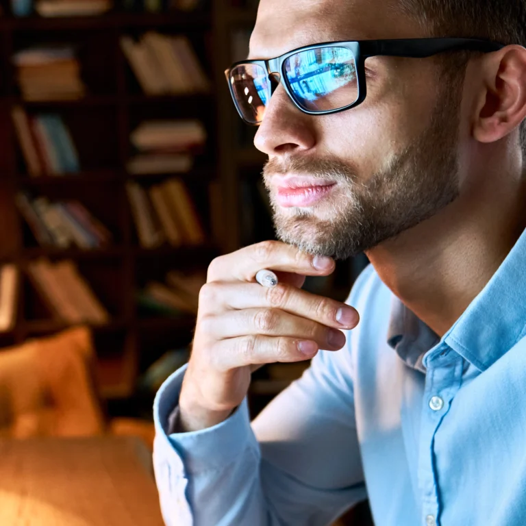 Man in glasses and light blue shirt sits indoors, resting his chin on his hand, looking thoughtfully into the distance with bookshelves in the background.