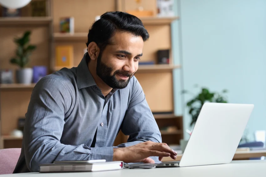 Man with a beard sitting at a desk, smiling while using a laptop. A notebook and pen are on the desk, with shelves and a plant in the background.