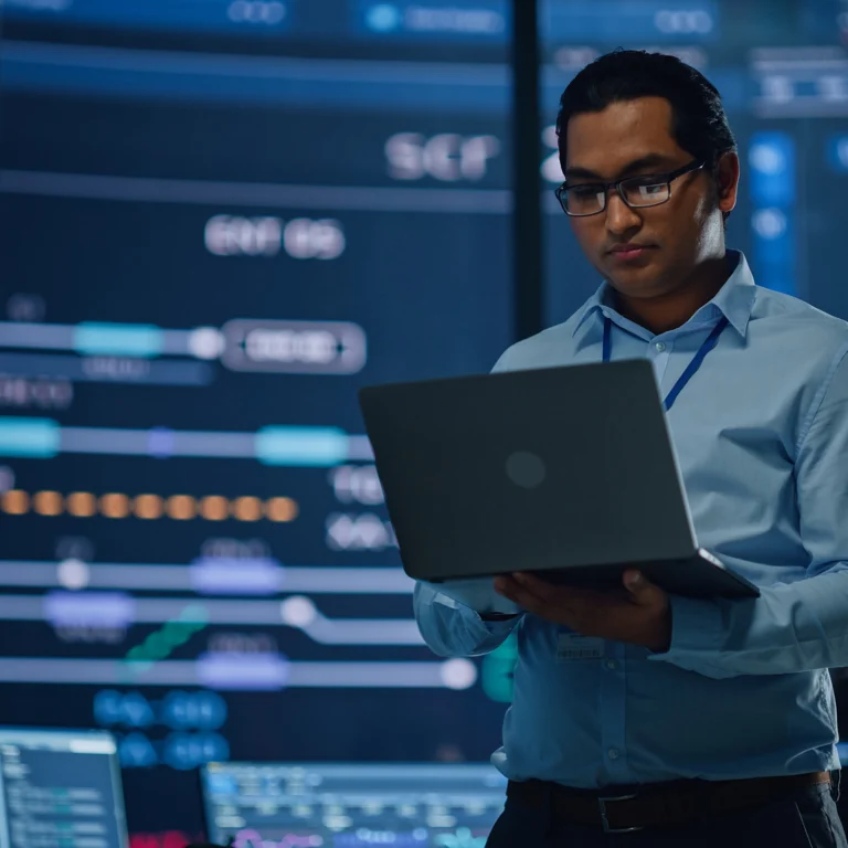 A person wearing glasses and a blue shirt works on a laptop in front of large digital screens displaying data and graphs.