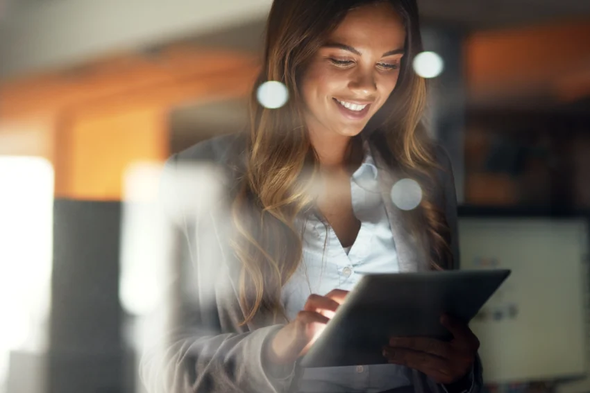 A woman stands indoors, smiling while using a tablet device. She has long hair and is dressed in business attire. The image has some light reflections in the foreground.