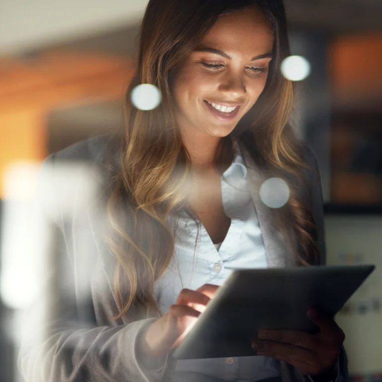 A woman in business attire is smiling while using a tablet, with soft lights and reflections visible in the foreground.