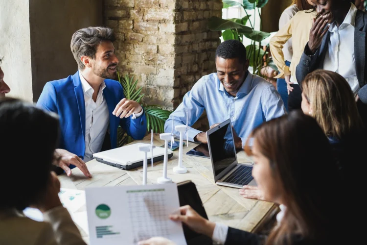 A group of people sit around a table with laptops and documents, having a discussion in a modern office setting.
