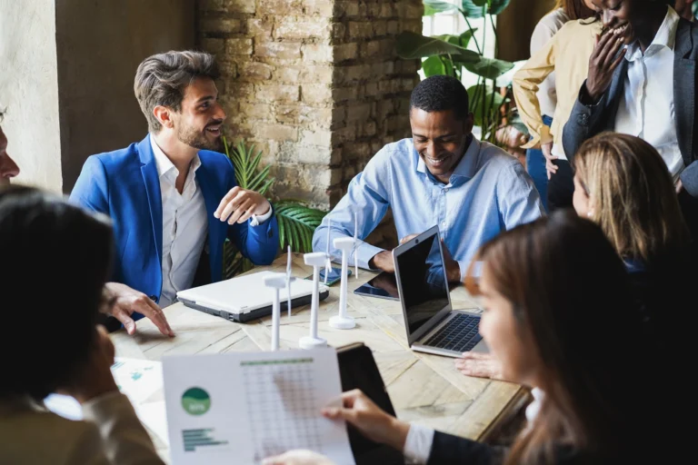 A group of people sit around a table with laptops and documents, having a discussion in a modern office setting.
