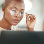 A person with short hair and glasses is looking intently at a laptop screen, adjusting their eyewear with one hand.
