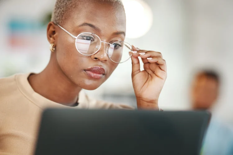 A person with short hair and glasses is looking intently at a laptop screen, adjusting their eyewear with one hand.