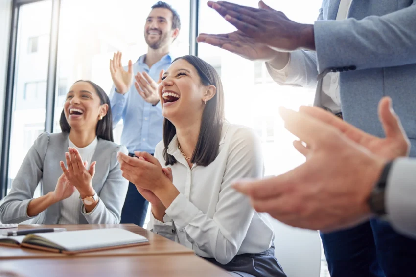 A group of professionals sitting and standing around a table, smiling and applauding in a bright office setting.