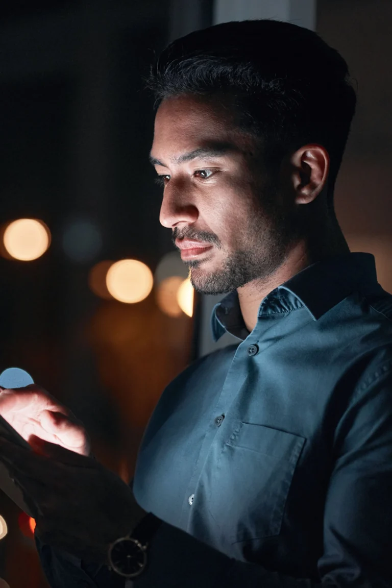 A man in a blue shirt uses a smartphone at night, illuminated by the screen, with blurred city lights in the background.