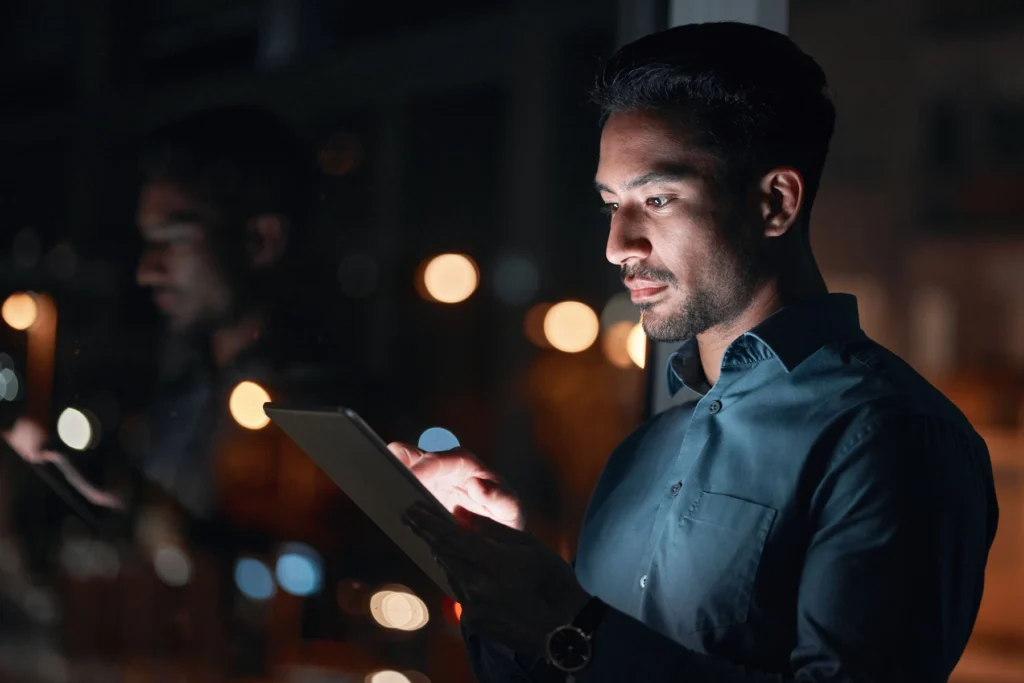 A man in a dark shirt uses a tablet at night, with city lights and his reflection visible in the window beside him.