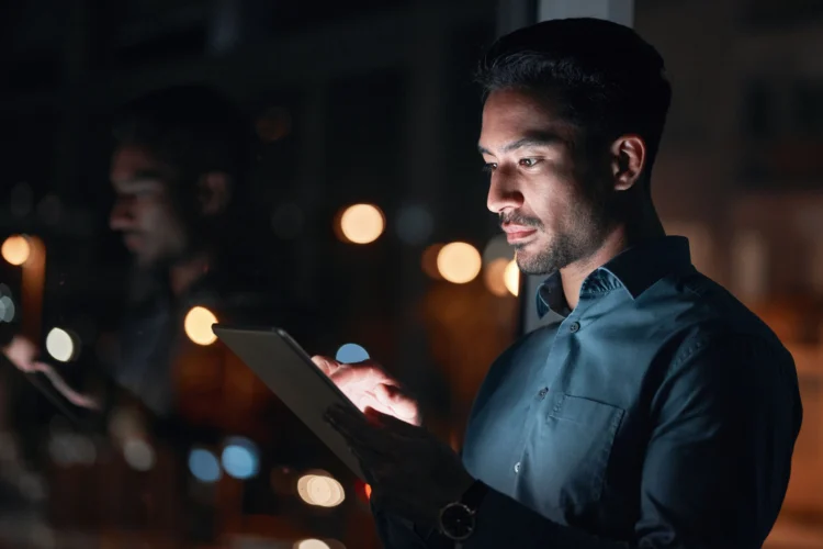 A man in a dark shirt uses a tablet at night, with city lights and his reflection visible in the window beside him.