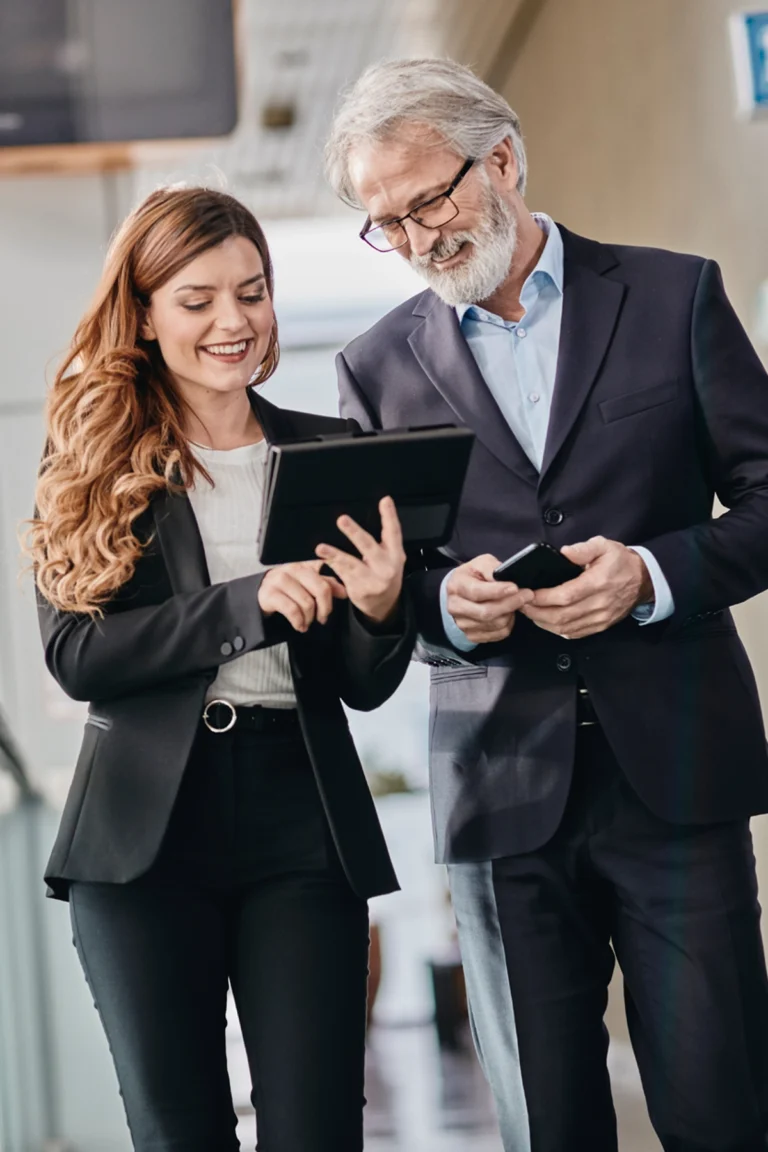 Two business professionals in suits stand together indoors, looking at a digital tablet and smartphone, appearing engaged in discussion.