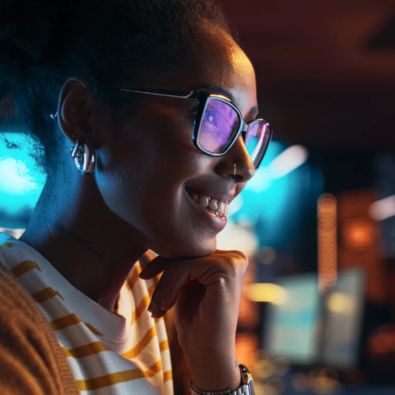 A person wearing glasses and a striped shirt smiles while looking at a computer screen in a dimly lit room.