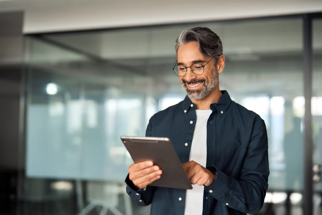 A man with glasses and gray hair uses a tablet while standing in a modern office with glass walls, smiling and looking at the screen.
