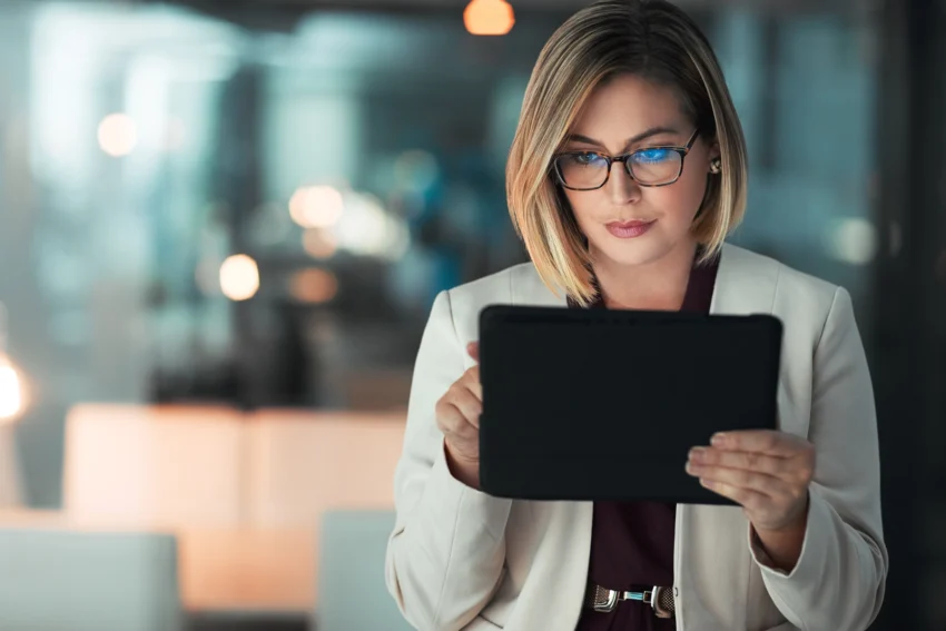 A woman in glasses and a blazer looks intently at a tablet in an office setting with blurred lights in the background.
