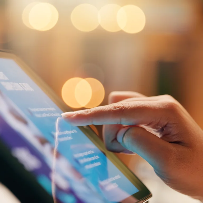 A person’s finger touches the screen of a digital tablet, with blurred lights in the background.