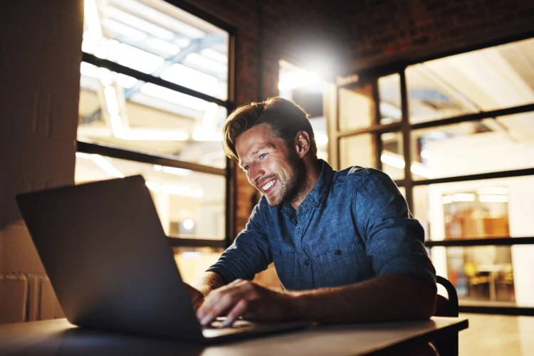 A man in a denim shirt sits at a desk, typing on a laptop and smiling, in a modern office with glass walls and exposed brick.
