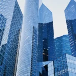 Tall modern glass skyscrapers reflecting each other against a clear blue sky in a city business district.