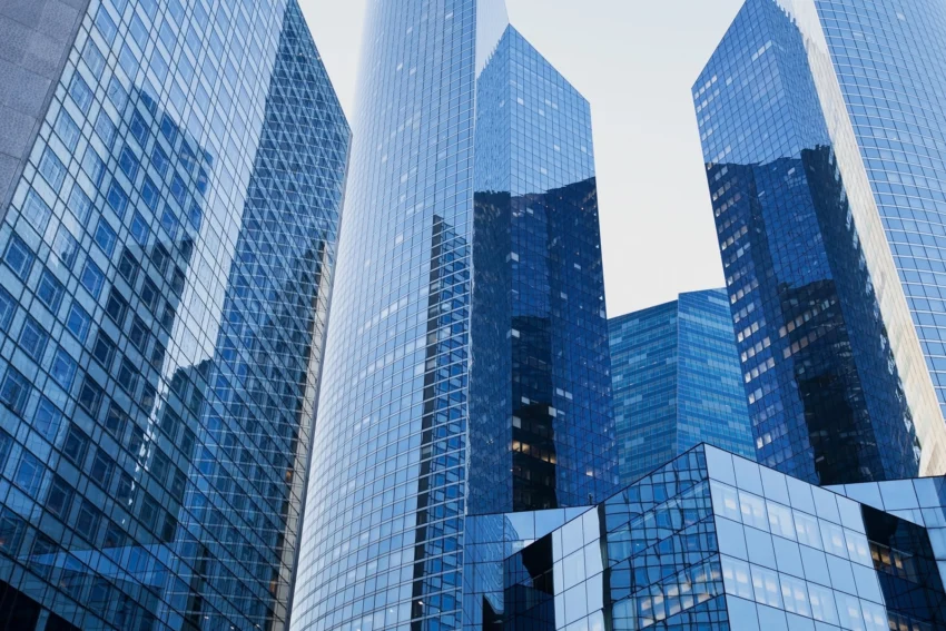 Tall modern glass skyscrapers reflecting each other against a clear blue sky in a city business district.