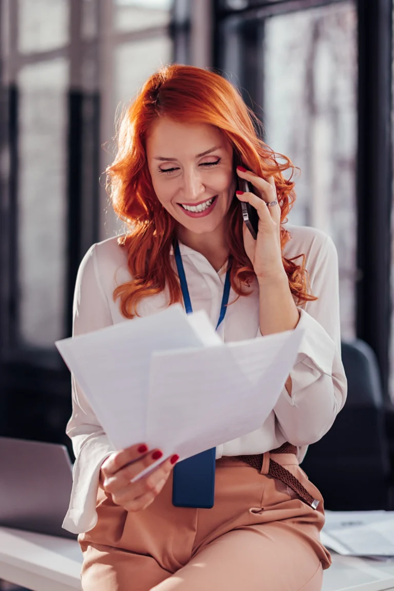 A woman with red hair, wearing a white blouse and beige pants, sits on a desk, smiling while talking on the phone and holding paperwork.