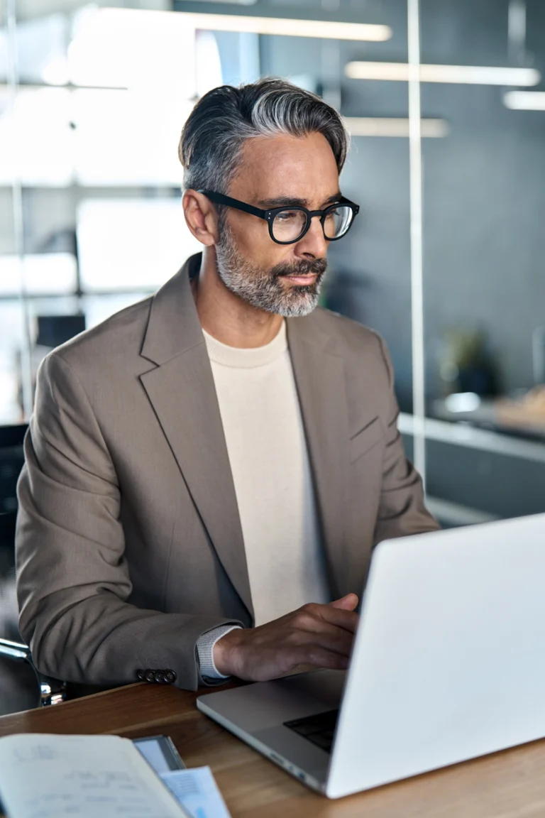 A man wearing glasses and a beige blazer sits at a desk working on a laptop in a modern office setting.