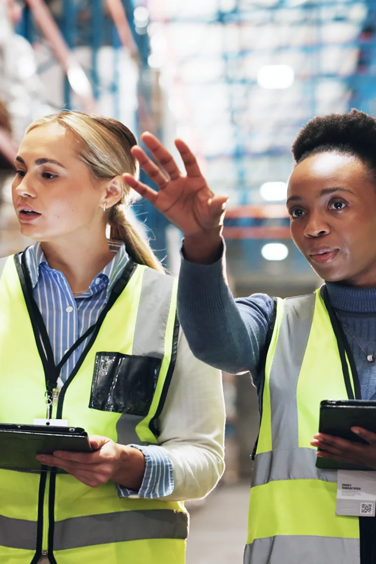 Two women in safety vests holding tablets stand in a warehouse, with one pointing and explaining something to the other.