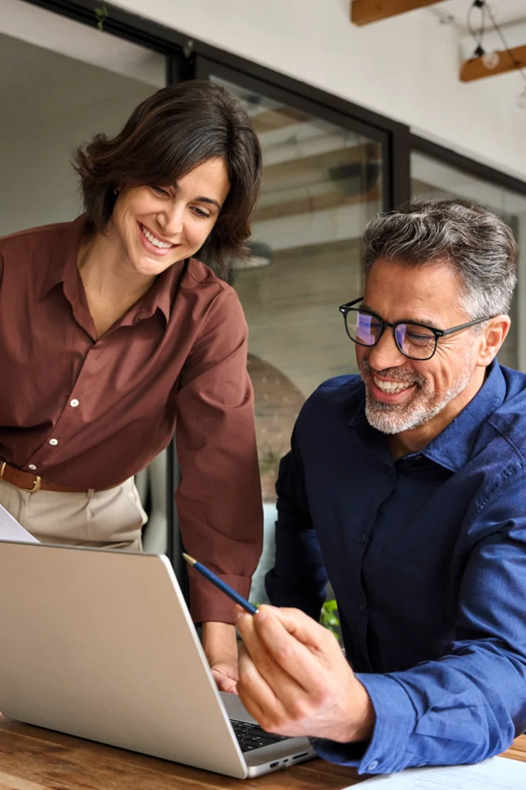 Two colleagues, one standing and one sitting, collaborate and smile while looking at a laptop on a desk in a modern office setting.