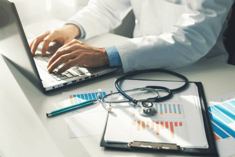 A person in a white coat types on a laptop at a desk with medical charts, a clipboard, a pen, and a stethoscope.