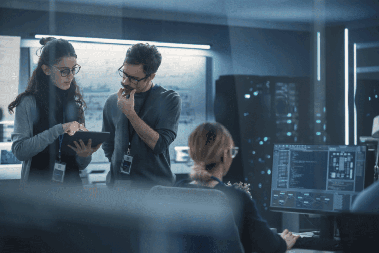 Three people in an office, two standing and discussing something on a tablet, one sitting at a desk working on a computer with code on-screen. Whiteboards and monitors in the background.