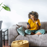 A person sitting cross-legged on a sofa, looking at a smartphone and smiling excitedly, with a large potted plant nearby.