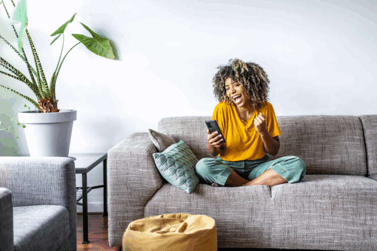 A person sitting cross-legged on a sofa, looking at a smartphone and smiling excitedly, with a large potted plant nearby.