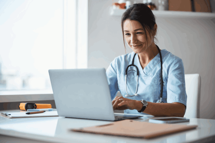 A healthcare professional in scrubs with a stethoscope works on a laptop at a desk in a well-lit office.